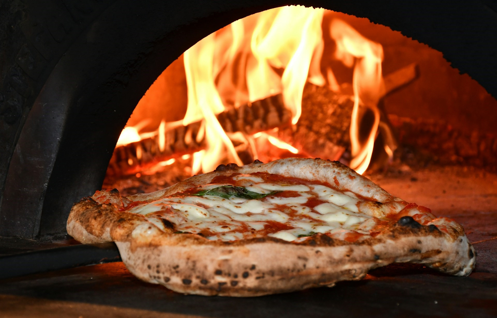 Pizza wordt uit een steenoven gehaald, met op de achtergrond vuurvlammen en houtstukken