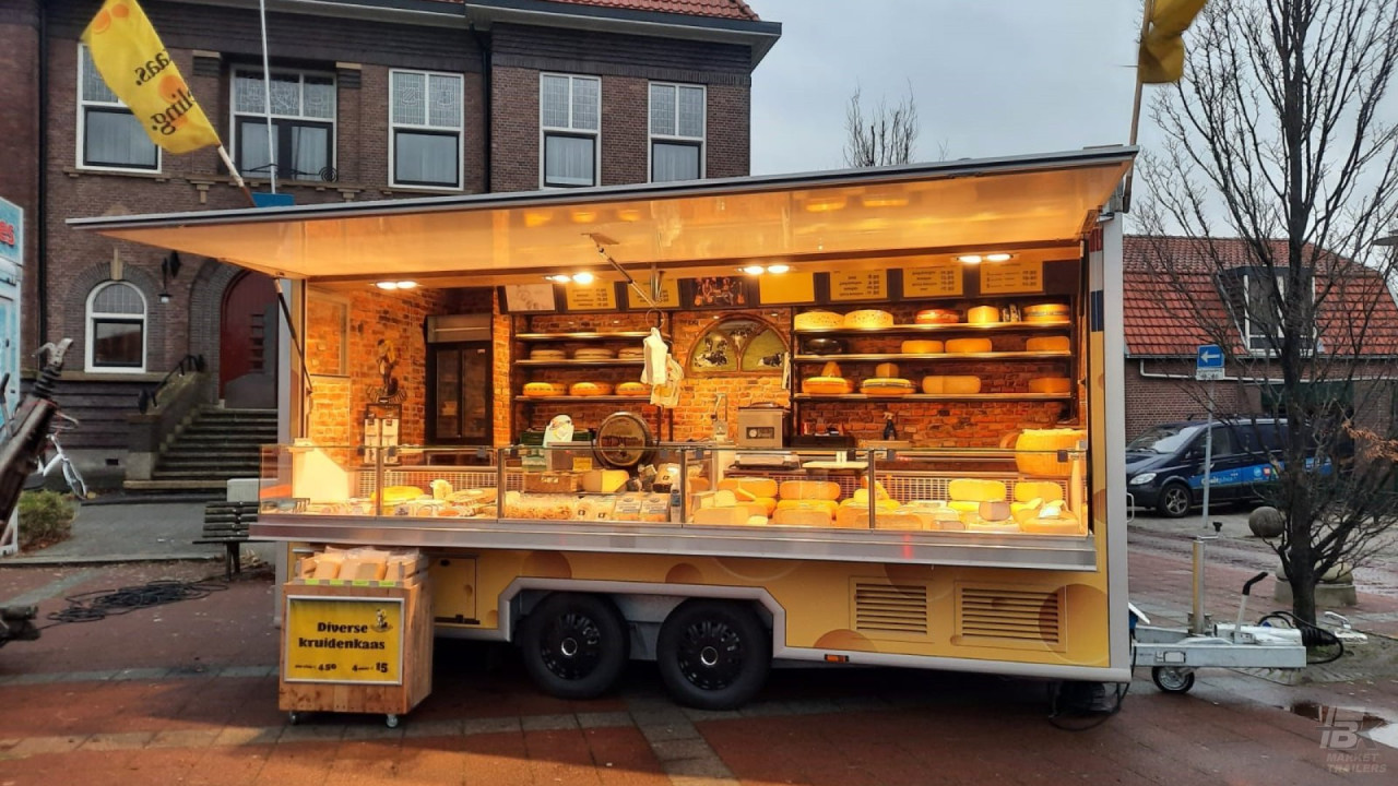 Zijaanzicht van een aanhangwagen voor kaas met geopende luifel, gevuld met het assortiment