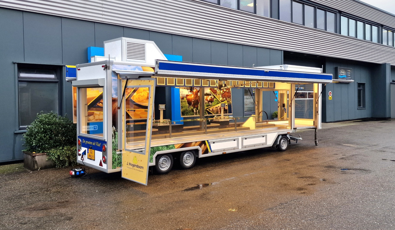 Zijaanzicht van een smalle aanhangwagen voor kaas met geopende luifel en zijbord